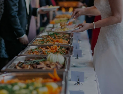 A catered table with a bride wearing a white dress and silver bracelet getting food.