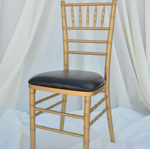 Gold colored chiavari chair with a black seat cushion in front of  a white backdrop.